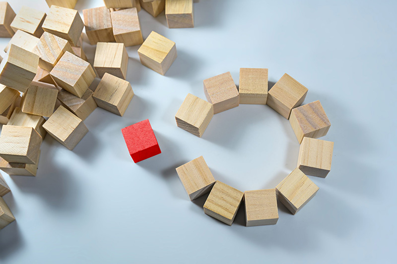 A group of plain wooden blocks forms a rough circle with an opening. A single red block is placed just outside the gap, standing out from the other blocks.